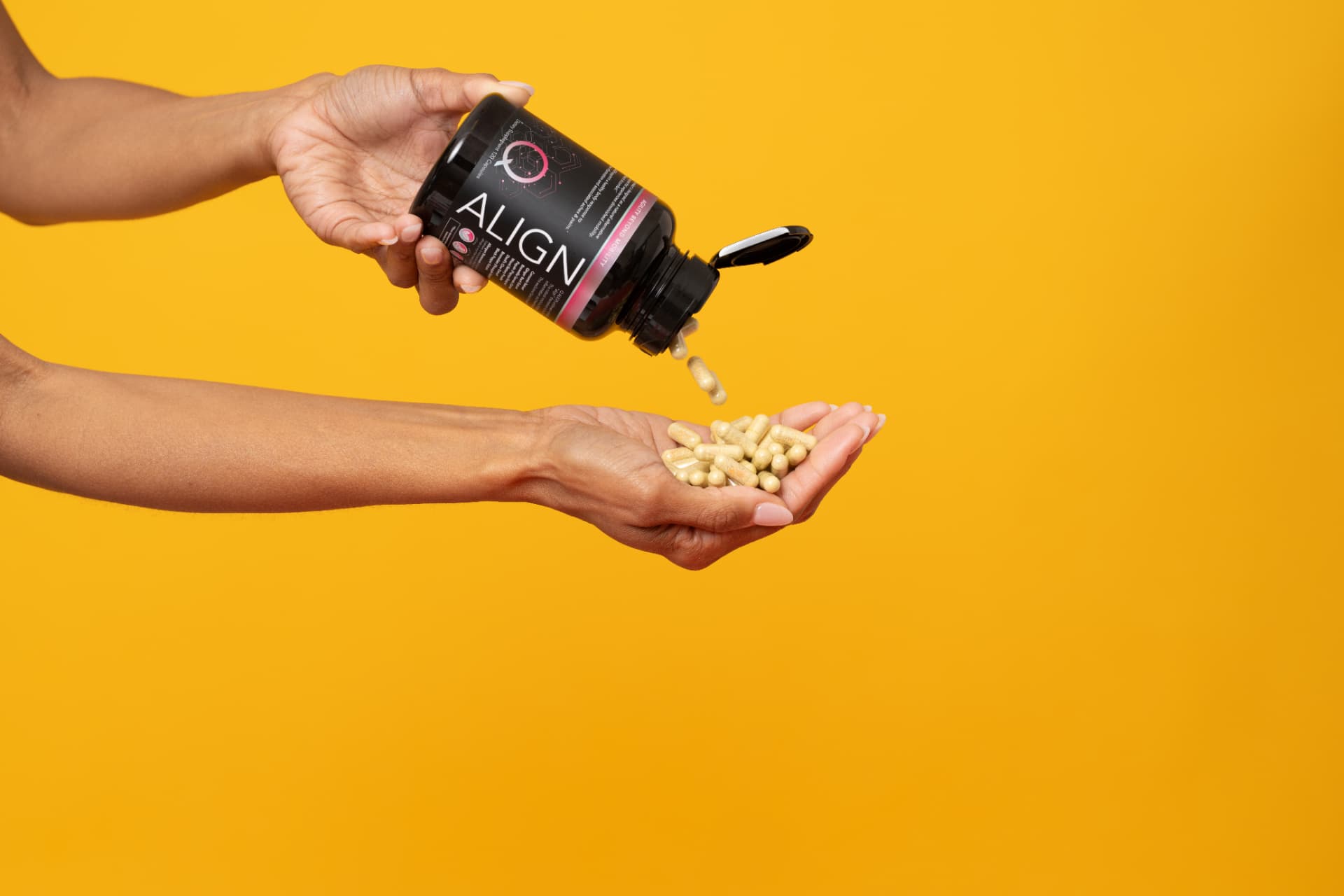 Hands pouring supplement capsules from a black bottle against a yellow background